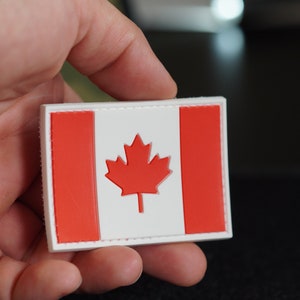 Puede incluir: Un parche rectangular con la bandera canadiense. El diseño presenta un fondo blanco con una hoja de arce roja en el centro, flanqueada por dos barras rojas verticales. El parche tiene un borde blanco.