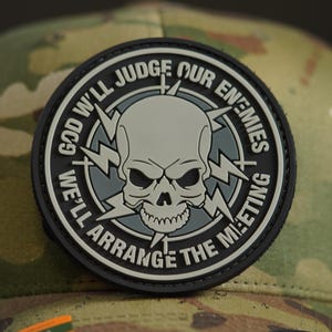 May include: A black and grey embroidered patch featuring a skull and crossbones design. The text surrounding the skull reads "God Will Judge Our Enemies We'll Arrange The Meeting".