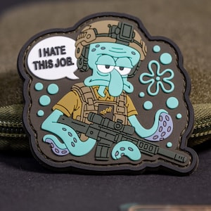 May include: A rubber morale patch featuring a light blue cartoon character wearing a tan tactical vest and helmet, holding a black rifle. A white speech bubble contains the black text I HATE THIS JOB. The design includes light blue bubbles.