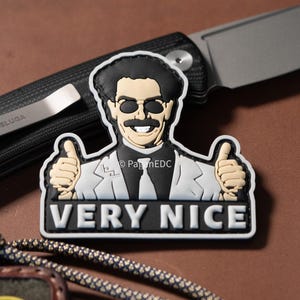 May include: A black, white, and tan patch featuring a cartoon character with sunglasses and a mustache giving a thumbs-up. The text "VERY NICE" is below the character. A knife is in the background.