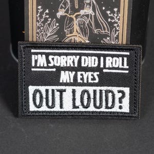 Puede incluir: Un parche rectangular bordado en negro con un borde blanco y texto blanco cosido que dice, I'M SORRY DID I ROLL MY EYES OUT LOUD? El parche presenta un estilo de fuente claro y negrita con una textura de tela estructurada y borde acabado.