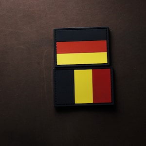 May include: Two black fabric patches with embroidered flags. The top patch features the German flag with black, red, and yellow horizontal stripes. The bottom patch features the Belgian flag with black, yellow, and red vertical stripes.