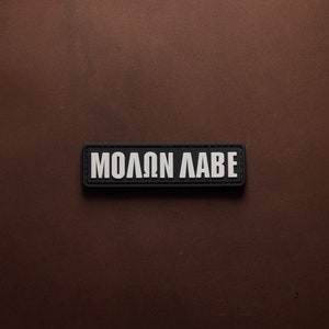 Puede incluir: Un parche negro con texto blanco que dice "MOLON LABE".