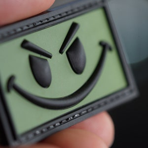 Può includere: Una toppa verde e nera con un viso sorridente con un'espressione maliziosa. La toppa ha un bordo nero ed è realizzata in un materiale testurizzato.