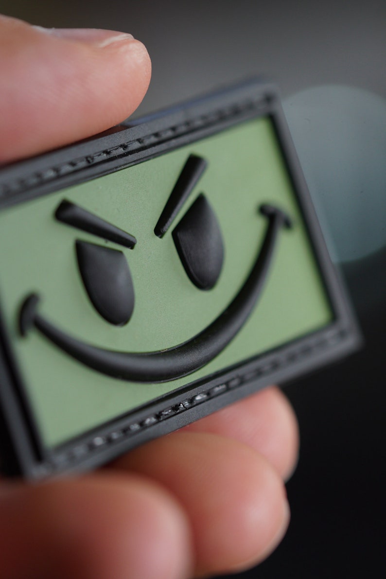 May include: A green and black patch with a smiling face with a mischievous expression. The patch has a black border and is made of a textured material.