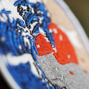 Op de afbeelding: Close-up van een geborduurde patch met een blauwe golf met een rode zon die erachter opkomt. De patch is rond en heeft een witte achtergrond.