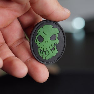 Puede incluir: Un parche de calavera negro y verde con un borde cosido. La calavera es de estilo de dibujos animados con una cara verde y ojos negros.