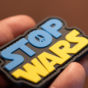 Peut inclure: Un patch noir avec les mots "STOP WARS" en lettres bleues et jaunes. Un symbole de paix se trouve au centre du mot "STOP".