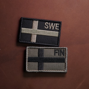 Puede incluir: Dos parches de tela con banderas bordadas. El parche superior es negro con una cruz blanca y el texto "SWE". El parche inferior es verde con una cruz negra y el texto "FIN".