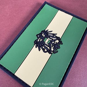 Peut inclure: Écusson rectangulaire avec un design à rayures vertes et beiges, orné d'un emblème noir au centre. Le patch a une bordure noire et le texte "PaganEDC" est visible dans le coin inférieur droit.