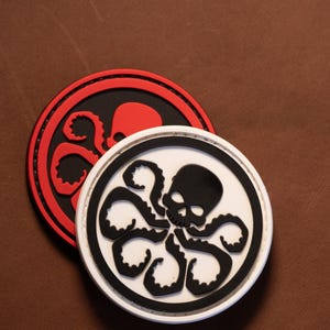 May include: Two circular patches featuring a raised graphic of a skull with octopus tentacles extending from the base. One patch has a white background with a black skull and tentacles, while the other has a red background with a black skull and tentacles.