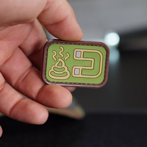 May include: A rectangular patch with a green background and a brown border. The patch features a cartoon image of a pile of poop with steam rising from it, and the number "5" in a stylized font. The patch is held by a hand.