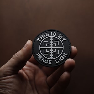 Puede incluir: Un parche negro y blanco con el texto "This is my peace sign" y un símbolo de paz estilizado.