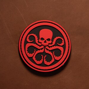 May include: A circular patch featuring a raised red skull and tentacle design set against a black background. The emblem is bordered by a thick red ring with visible stitching along the edge, creating a distinct contrast between the red and black.