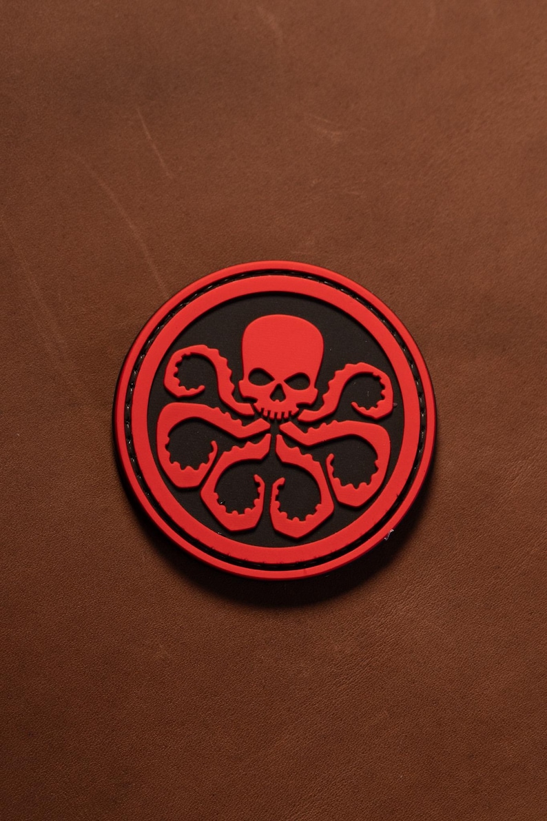 May include: A circular patch featuring a raised red skull and tentacle design set against a black background. The emblem is bordered by a thick red ring with visible stitching along the edge, creating a distinct contrast between the red and black.