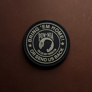 Può includere: Un cerotto rotondo nero e marrone con il testo "BRING 'EM HOME!" e "POW-MIA" con una silhouette di una testa di soldato. Il testo "OR SEND US BACK" è sotto la silhouette e il testo "YOU ARE NOT FORGOTTEN" è sotto quello.