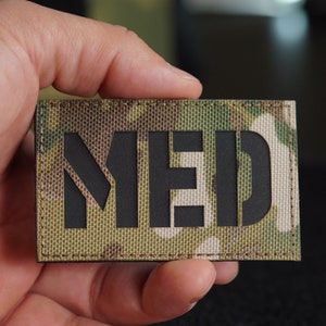 May include: A black patch with the word "MED" in white letters on a camouflage background.