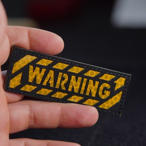 Könnte beinhalten: Ein rechteckiger Aufnäher mit schwarzem Hintergrund und gelbem Warnmuster, der das Wort WARNING in fetten Blockbuchstaben zeigt. Die gelben Bereiche haben eine sichtbare hexagonale Wabenstruktur und der Aufnäher hat einen genähten Rand.