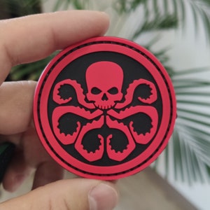 May include: A circular patch featuring a raised red graphic of a skull with six tentacles extending downwards against a solid black background. The design is enclosed within a thick red border with visible stitching along the outer edge of the circle.