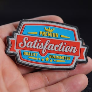 Puede incluir: Un parche gris, azul, rojo y amarillo con un borde cosido. El parche tiene una corona y el texto "Premium Satisfaction Quality Guaranteed" con cinco estrellas.
