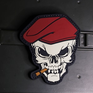 May include: A white skull with a red beret and a cigarette in its mouth. The skull is outlined in black and has a stitched border.