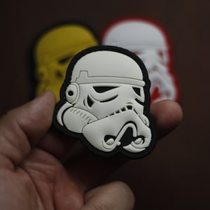 Peut inclure: Un patch en plastique blanc et noir représentant un casque de Stormtrooper de Star Wars. Le patch est tenu dans une main.