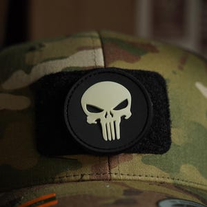 May include: A black and green camouflage baseball cap with a black velcro patch featuring a white glow-in-the-dark skull graphic.