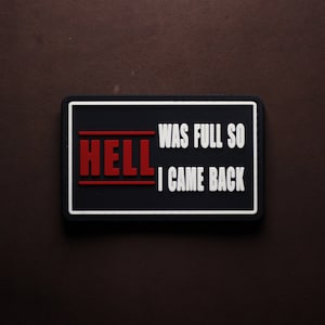 Puede incluir: Un parche rectangular negro con el texto blanco: «HELL WAS FULL SO I CAME BACK». La palabra «HELL» está en rojo. El parche tiene un borde blanco y está sobre un fondo marrón.
