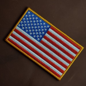 Op de afbeelding: Een klein geborduurd Amerikaans vlagpatch met een gouden rand. De vlag is rood, wit en blauw.
