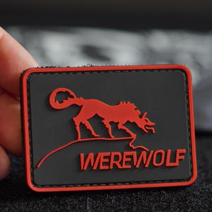 Può includere: Una toppa ricamata nera e rossa con una silhouette di lupo mannaro rossa e la parola "WEREWOLF" in rosso.