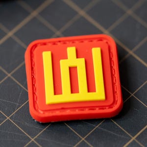 May include: A red and yellow patch with a stylized symbol of a building. The patch is made of a soft, flexible material and has a stitched border.
