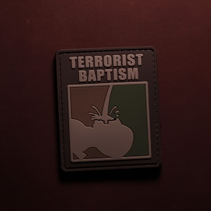 May include: A rectangular patch with the words "TERRORIST BAPTISM" in white block letters. The patch features a graphic of a person being baptized with brown and green sections. The patch has a dark gray border.