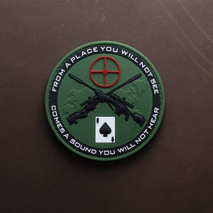 Può includere: Un cerotto rotondo verde e nero con un asso di picche bianco, due fucili incrociati e il testo "From a place you will not see comes a sound you will not hear".