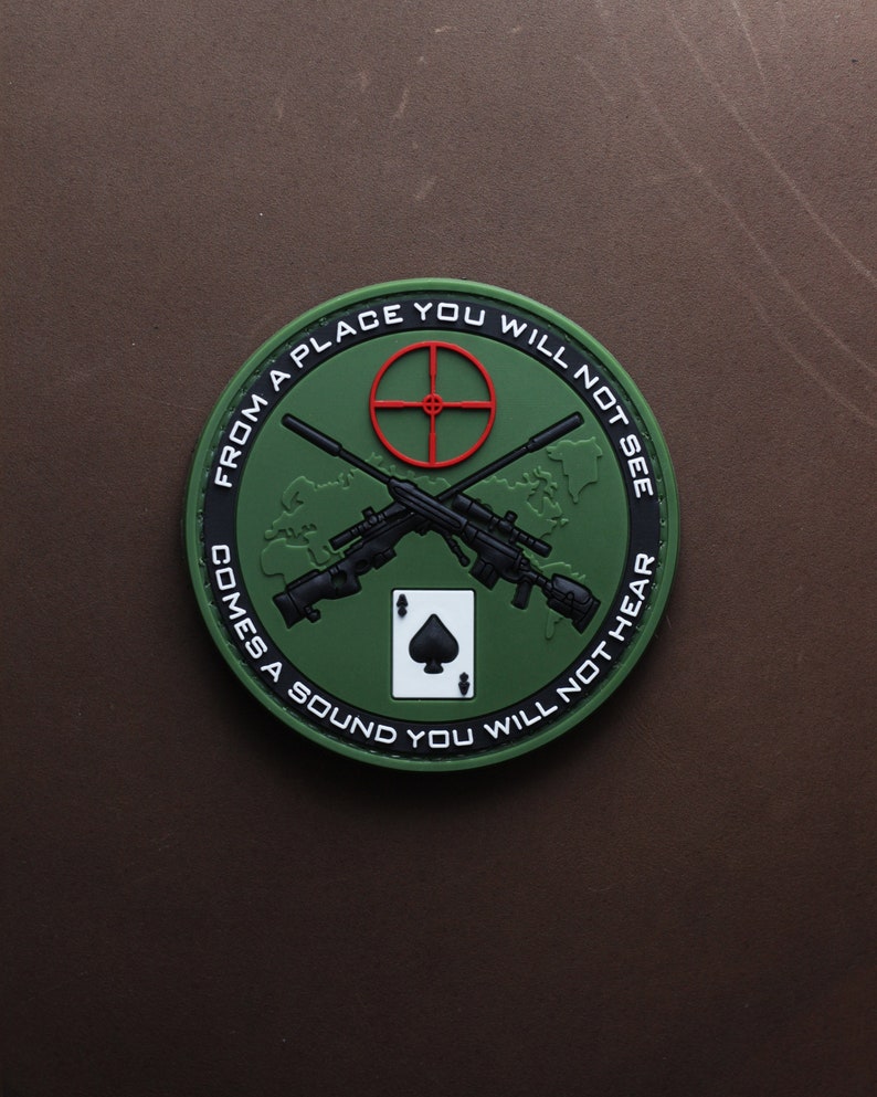 May include: Green and black round patch with a white ace of spades, two crossed rifles, and the text "From a place you will not see comes a sound you will not hear".