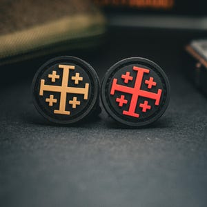 Może przedstawiać: Dwie czarne, okrągłe naszywki z motywem krzyża jerozolimskiego. Jedna naszywka ma złoty krzyż, druga czerwony. Naszywki leżą na ciemnej powierzchni.