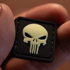 Può includere: Una toppa nera con un grafico di teschio bianco che brilla al buio. Il teschio ha un'espressione minacciosa con una mascella prominente e un naso appuntito. La toppa è cucita con filo nero attorno ai bordi.