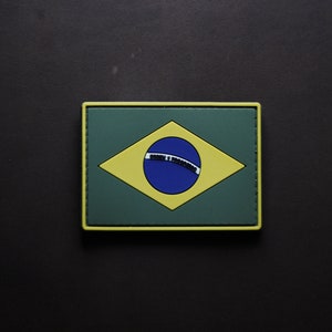 Puede incluir: Un parche bordado verde, amarillo y azul con la bandera de Brasil. El parche tiene un diamante amarillo con un círculo azul en el centro y una banda blanca a través del círculo. El diamante está sobre un fondo verde.