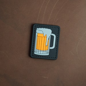 Op de afbeelding: Een zwart-witte patch met een cartoon afbeelding van een bierglas met een wit schuimkopje en oranje bier erin.