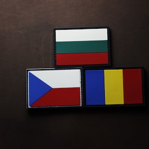 Puede incluir: Tres parches bordados con las banderas de Bulgaria, República Checa y Rumanía. Los parches son rectangulares y tienen bordes negros. La bandera búlgara es blanca, verde y roja. La bandera checa es blanca, azul y roja. La bandera rumana es azul, amarilla y roja.