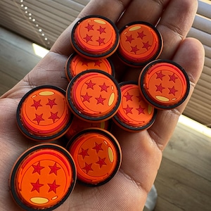 Può includere: Una collezione di ciondoli circolari arancioni e neri, ognuno con un diverso numero di stelle rosse. I ciondoli hanno una finitura lucida e sono tenuti in una mano, con una superficie di legno sullo sfondo.