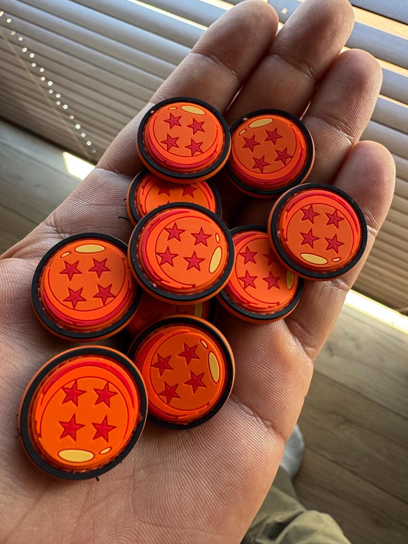 May include: A collection of circular orange shoe charms, each featuring a black border and four red star icons arranged in a diamond pattern, with a small yellow highlight detail on the surface of each individual round accessory piece.