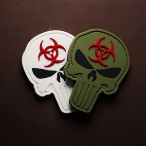 May include: Two skull patches, one white and one olive green, each featuring a red biohazard symbol. The skulls have black eye sockets and a stylized jawline. The patches are likely for tactical gear or apparel.