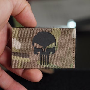 May include: A black and white Punisher skull patch on a camouflage fabric background.