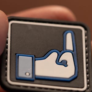 May include: A black and white patch with a blue outline of a Facebook thumbs up hand giving the middle finger.