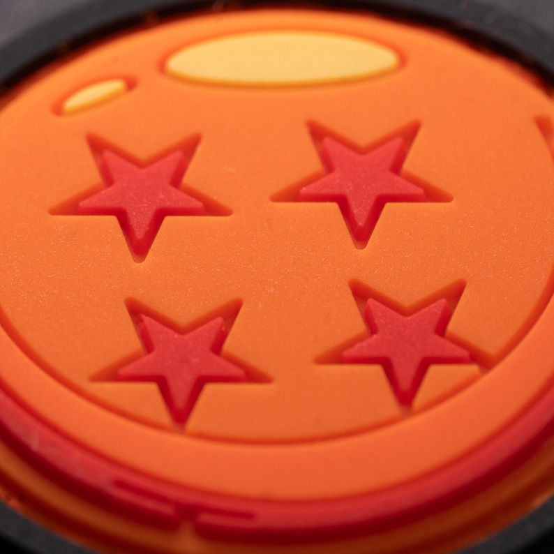 May include: A circular orange charm featuring four recessed red stars arranged in a square pattern. The surface has a smooth, matte texture with a light yellow oval highlight near the top edge, all encased within a thin black circular border.