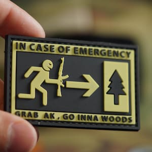 Pode incluir: Um remendo preto e amarelo com o texto "IN CASE OF EMERGENCY" e uma figura de pau correndo para a direita com um rifle, uma seta apontando para a direita e um pinheiro. O texto abaixo da imagem diz "GRAB AK, GO INNA WOODS".