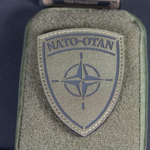 Peut inclure: Une pochette rectangulaire zippée vert olive avec un écusson en forme de bouclier centré sur le devant. L'écusson est vert olive avec une bordure gris foncé et le texte NATO-OTAN au-dessus d'un emblème en étoile à quatre branches.