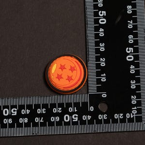 May include: A circular orange charm featuring four red stars and a yellow highlight, designed to resemble a four-star dragon ball. The charm is positioned next to a black L-shaped ruler, showing a diameter of approximately 1.25 inches.