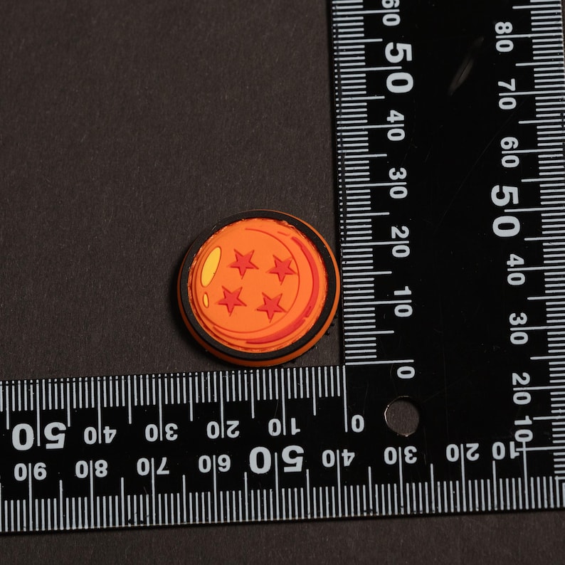 May include: A circular orange charm featuring four red stars and a yellow highlight, designed to resemble a four-star dragon ball. The charm is positioned next to a black L-shaped ruler, showing a diameter of approximately 1.25 inches.