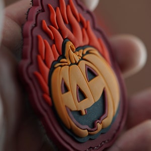 Può includere: Una toppa di Halloween rossa, arancione e nera con una zucca in fiamme. La toppa ha un bordo cucito ed è progettata per essere cucita su vestiti o una borsa.